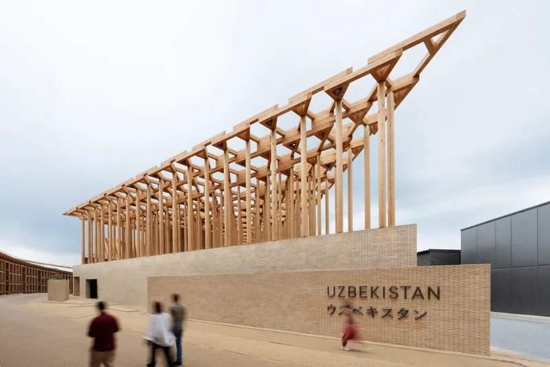 Diagonal timber roof structure rising above a beige brick facade with visitors walking past on an overcast day