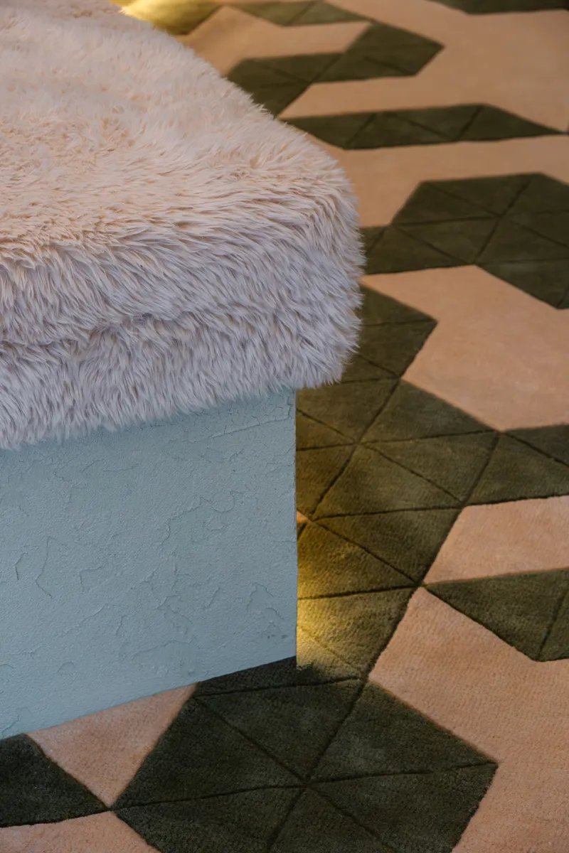 Detail of fur-upholstered bench edge meeting geometric carpet tiles in olive and pink tones