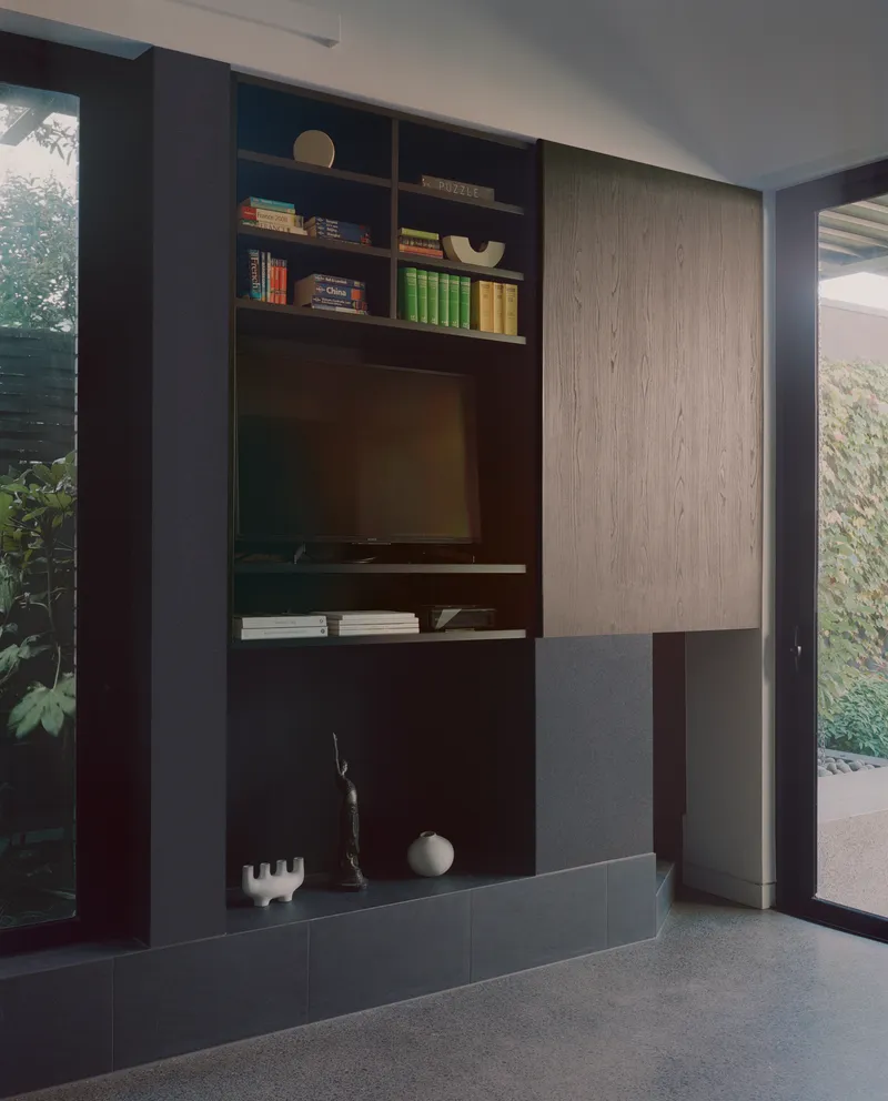 Built-in media unit with dark shelving and light oak cabinetry beside floor-to-ceiling glazing facing a garden