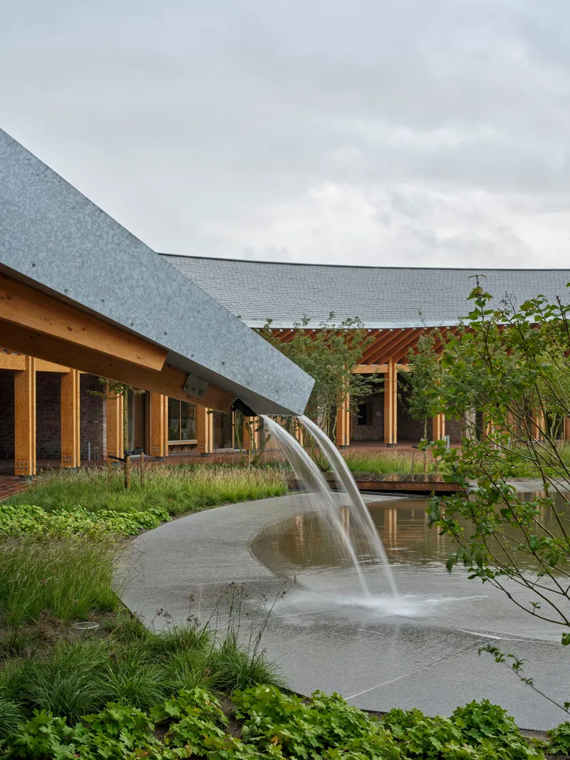 Courtyard water fountain with curved spout beside planted beds and overcast sky