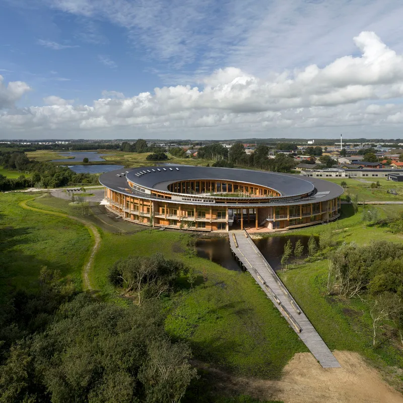 Aerial view of the ring-shaped structure with dark roof and timber colonnade approached by a wooden boardwalk