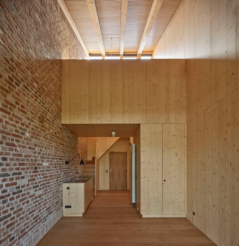 Double-height room with exposed brick wall, plywood volume, and slatted timber ceiling with clerestory windows
