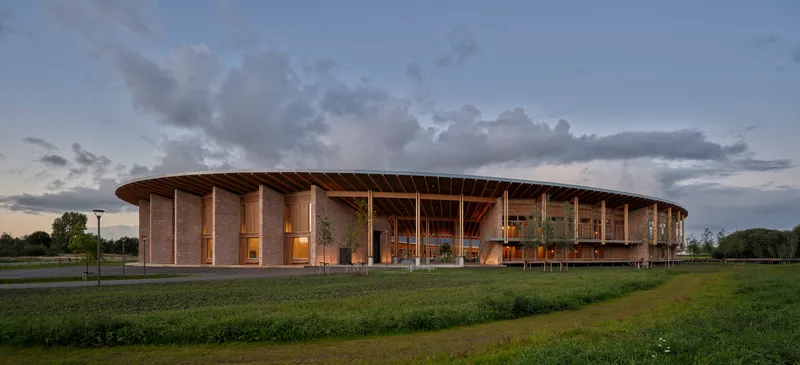 Circular colonnade with timber roof structure and brick piers across an open field at twilight