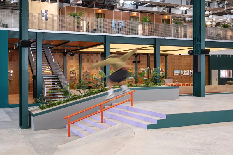 Double-height atrium with green steel columns, concrete skate ramp and a blurred skateboarder in motion