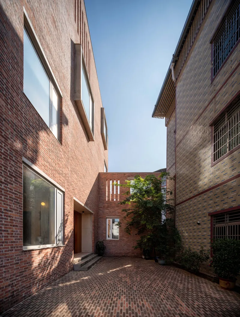 Brick courtyard between masonry volumes with planted tree under late afternoon light