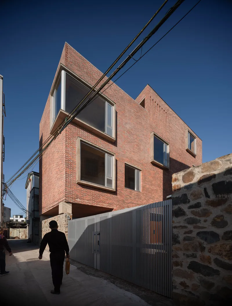 Street view of brick facade with asymmetric window openings and corrugated metal fence under clear blue sky