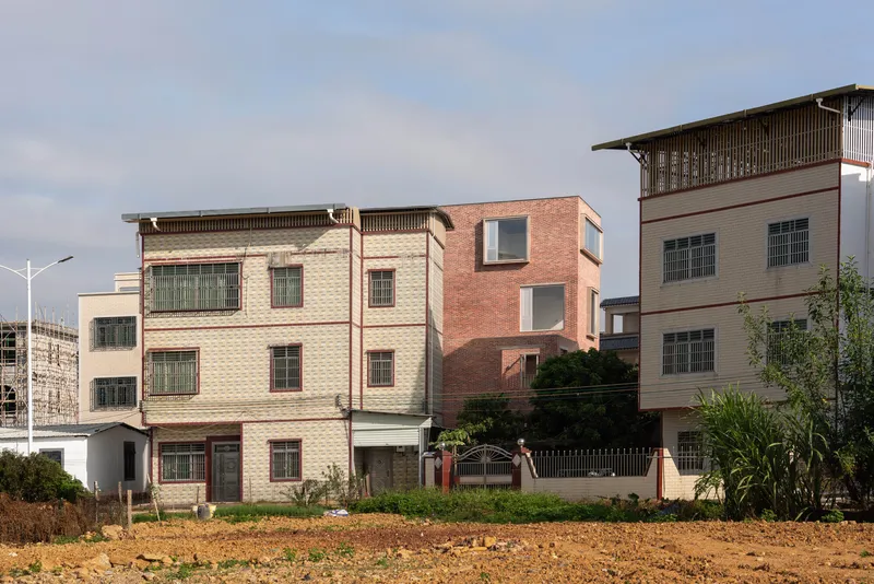 Stacked brick volumes with recessed windows alongside tiled neighboring structures under an overcast sky