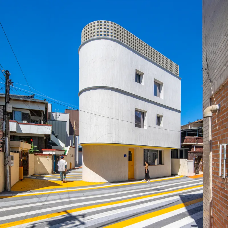 Three-story cylindrical volume with white facade, lattice rooftop parapet, and yellow-striped crosswalk extending toward the entrance
