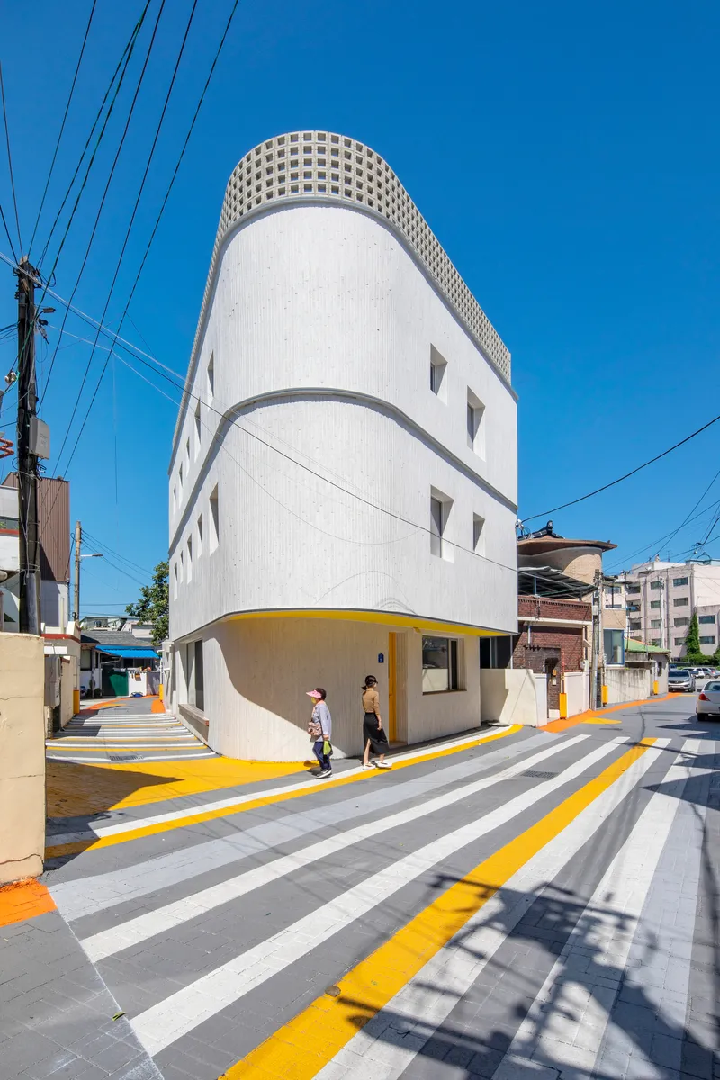 Corner view of the rounded building with lattice crown, white ribbed cladding, and yellow crosswalk under overhead power lines
