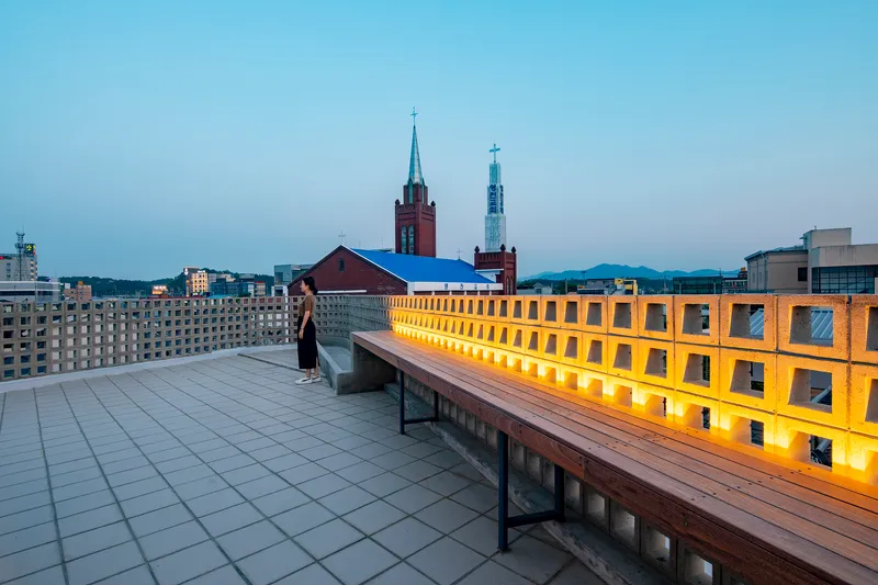 Rooftop terrace with timber bench and backlit perforated parapet facing church spires at twilight