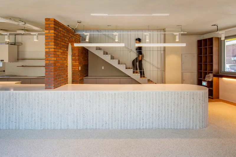 Open-plan interior with ribbed counter, glass-enclosed staircase, and exposed brick wall under fluorescent lighting
