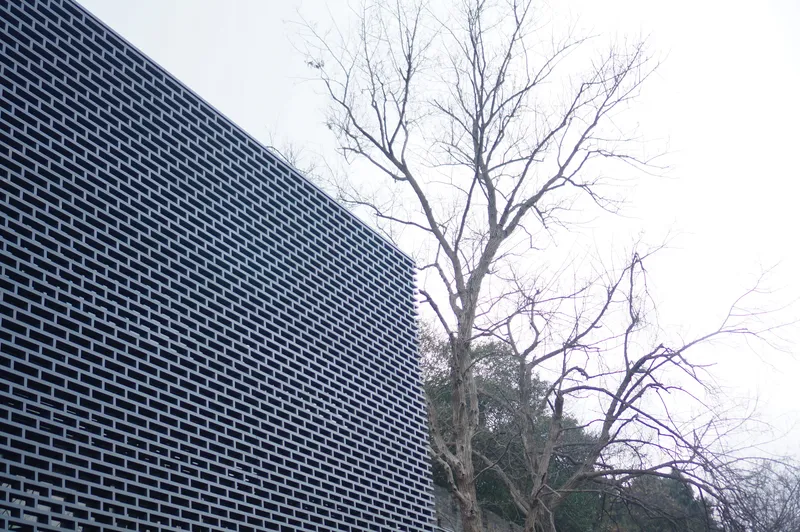 Perforated metal facade alongside bare winter tree branches against overcast sky