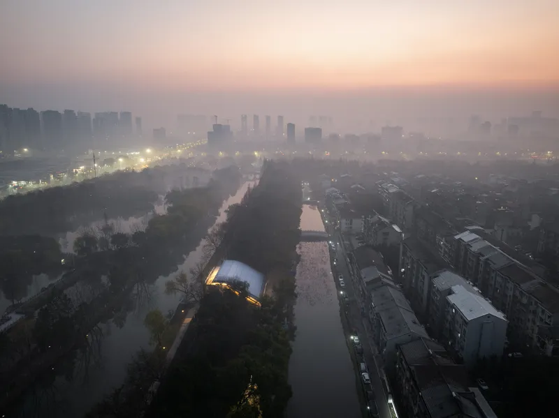 Aerial view of illuminated glass pavilion beside canal at dawn with heavy fog