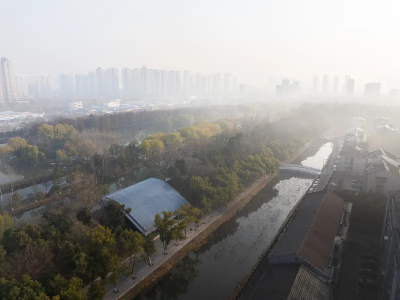 Aerial view of the arched metal roof nestled among autumn trees beside a canal and distant towers in heavy fog