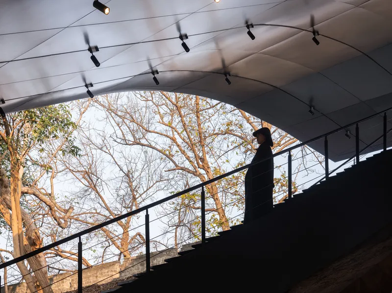 Figure ascending the interior staircase beneath a curved ceiling with track lighting and bare tree branches visible beyond