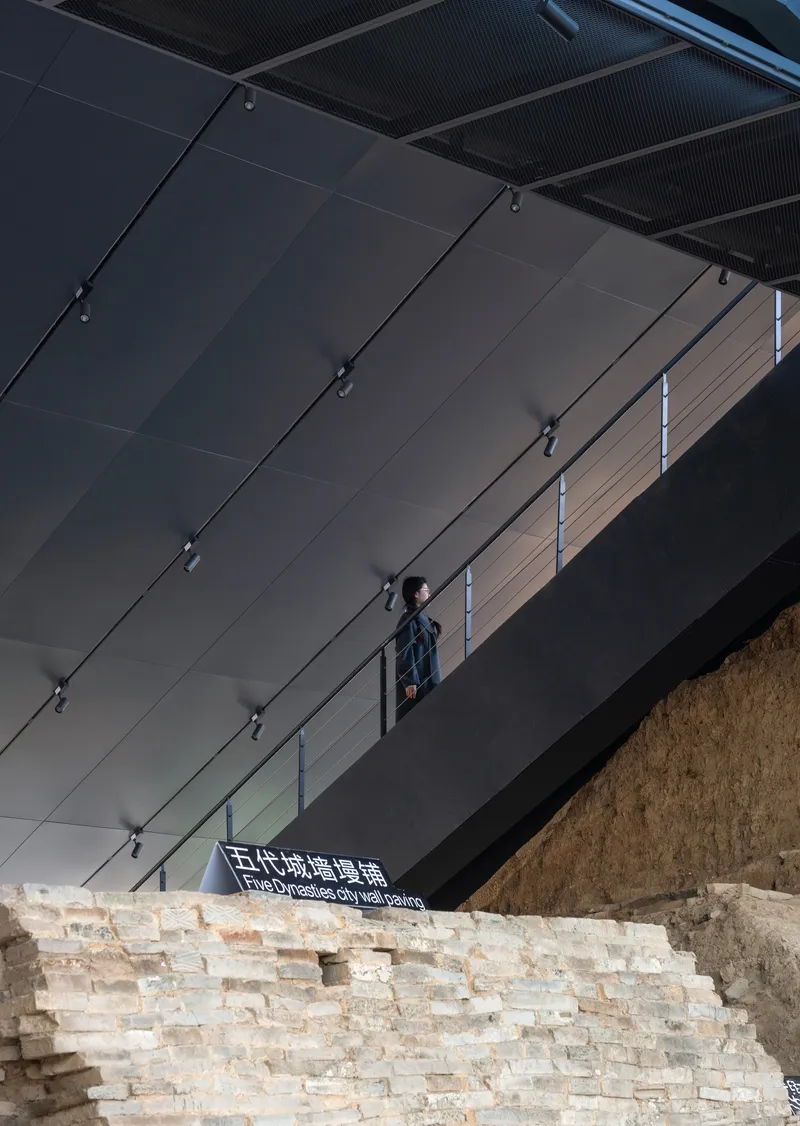 Person standing on the upper gallery above a reconstructed cream brick wall beneath a dark ceiling with track lighting