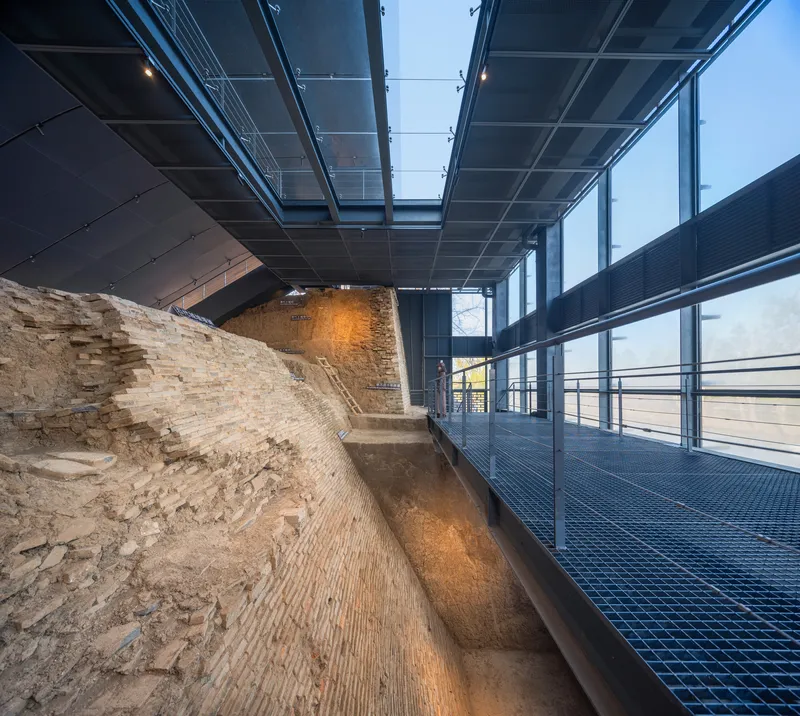 Interior walkway with glass roof beams and metal grating floor revealing the excavated rampart layers below