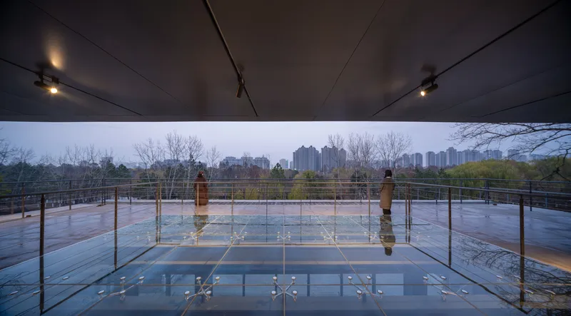 Glass floor terrace with steel railings reflecting bare trees and overcast sky