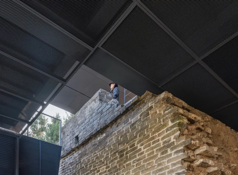 Visitor standing atop preserved brick wall remnant under perforated metal ceiling panels