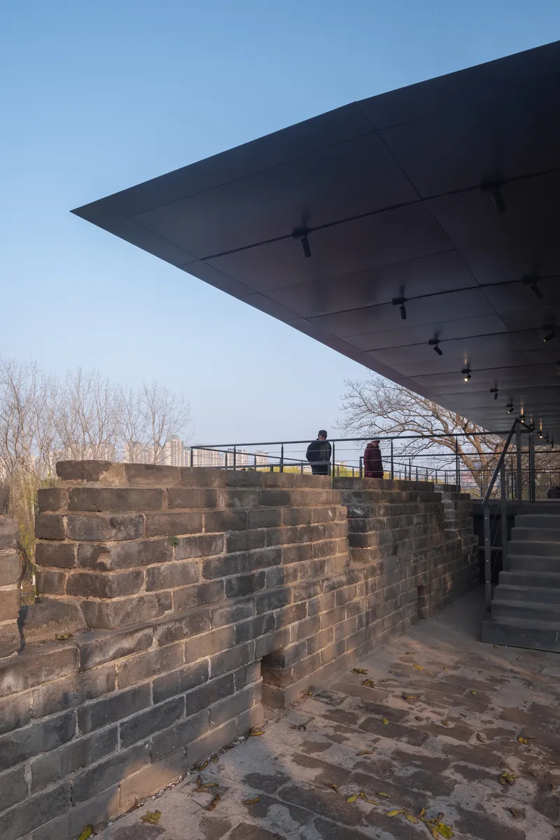 Cantilevered roof with track lighting extends over a historic brick wall path with two visitors at dusk