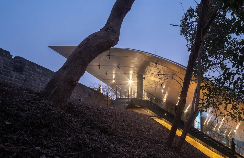Illuminated curved canopy shelters a glowing walkway beside an earthen slope with leaning trees at dusk