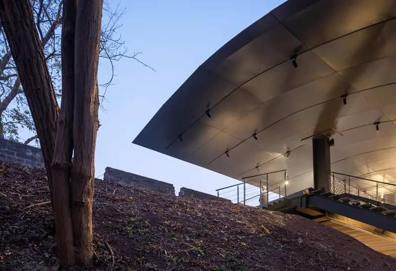 Underside of angular metal roof with track lighting hovers above sloped ground and tree trunks at twilight