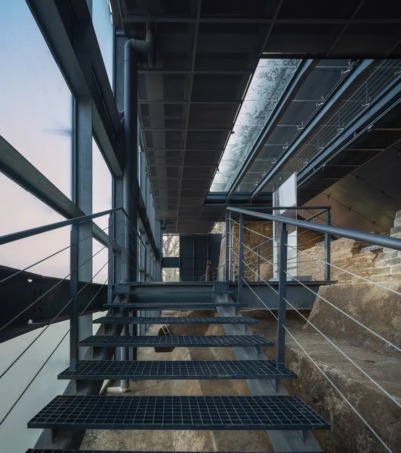 Metal mesh staircase descending between protective canopy and exposed archaeological brickwork