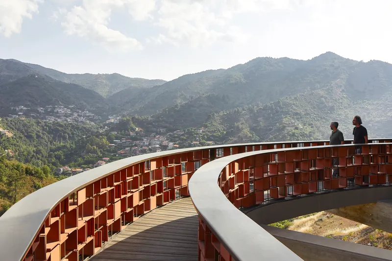 Curved terracotta screen wall with two figures silhouetted against distant mountain ridges