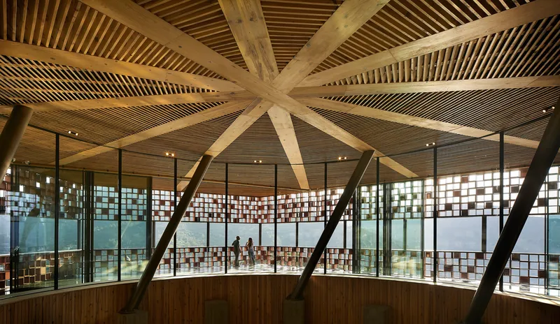 Interior view of radial timber beams converging at the ceiling center above glass-walled galleries with visitors