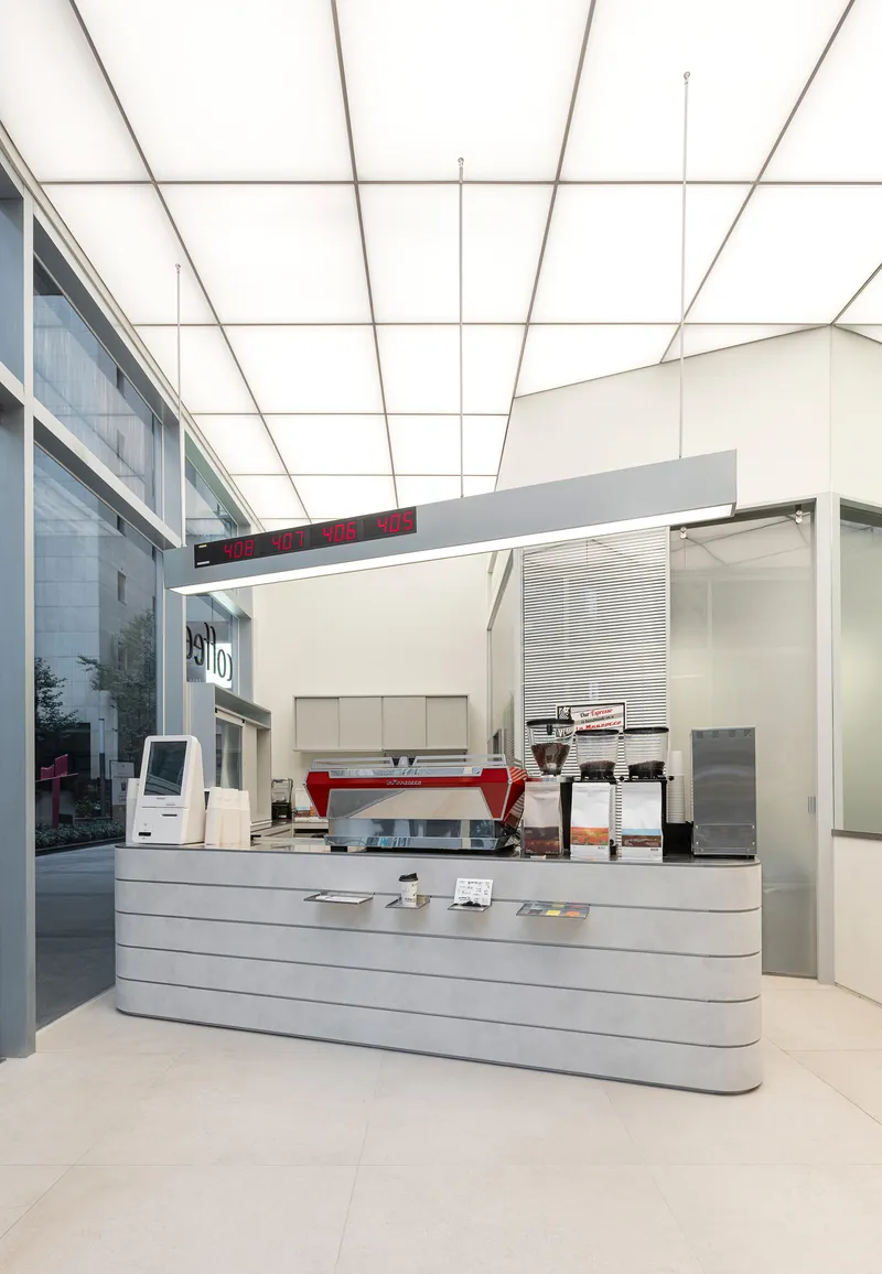 Red espresso machine on ribbed counter beneath translucent panel ceiling and glazed courtyard