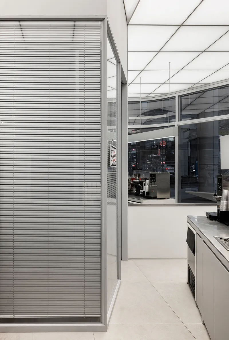 Interior corridor with horizontal metal blinds alongside glass wall under geometric ceiling grid