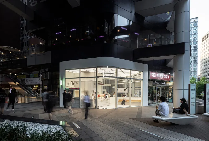 Glass storefront pavilion beneath a raised plaza structure at dusk with pedestrians passing by