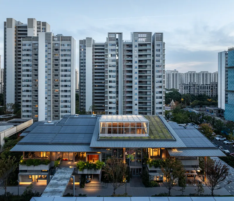Aerial view of low-rise timber structure with roof terrace surrounded by residential towers at dusk