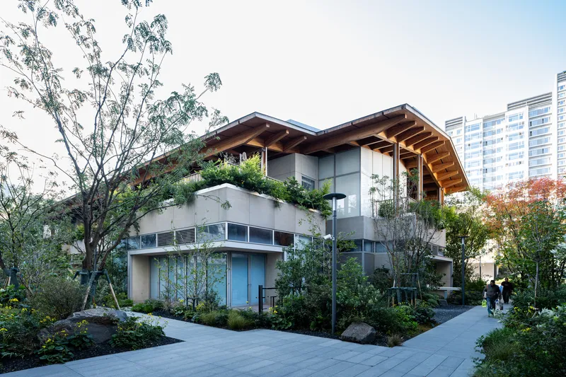 Two-story structure with planted roof terraces and exposed timber roof beams surrounded by trees and residential towers