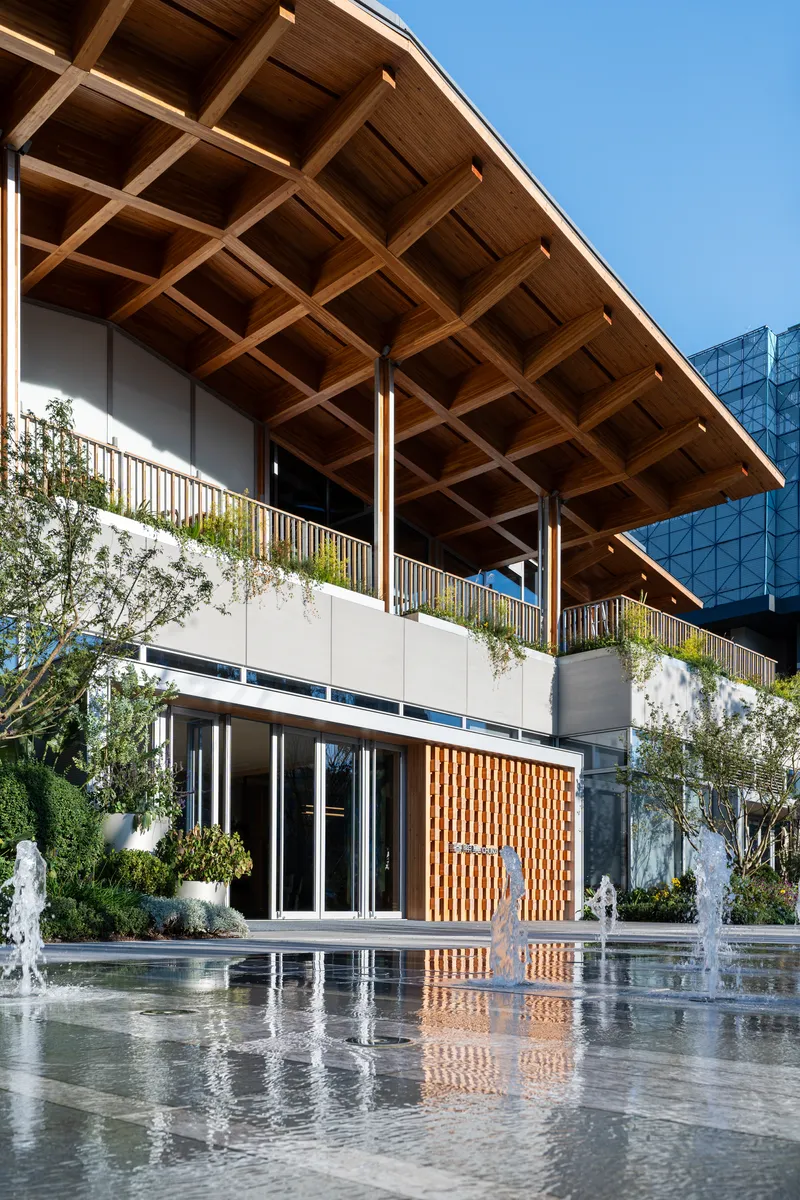 Timber coffered roof overhang above planted balcony reflected in shallow water feature with active fountains