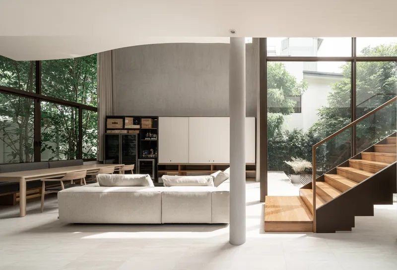 Living area with timber staircase, cylindrical columns, and floor-to-ceiling glazing overlooking greenery