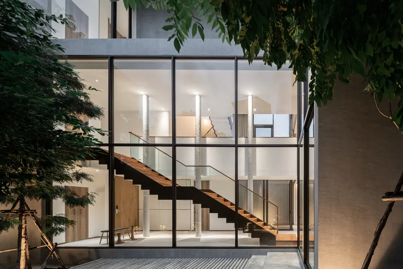 Courtyard view at twilight showing illuminated staircase through gridded glass facade surrounded by trees