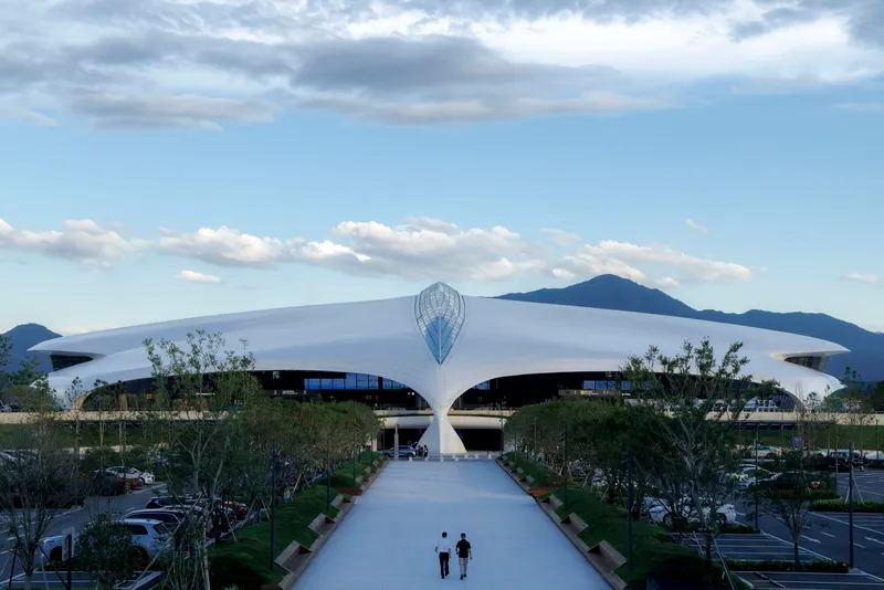 Two visitors walking the central arrival pathway toward the white curving roof canopy with mountains in the background