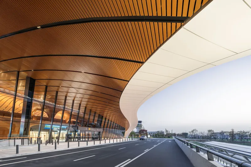 Curving timber-slat ceiling above the departure roadway and glazed entry facade at dusk