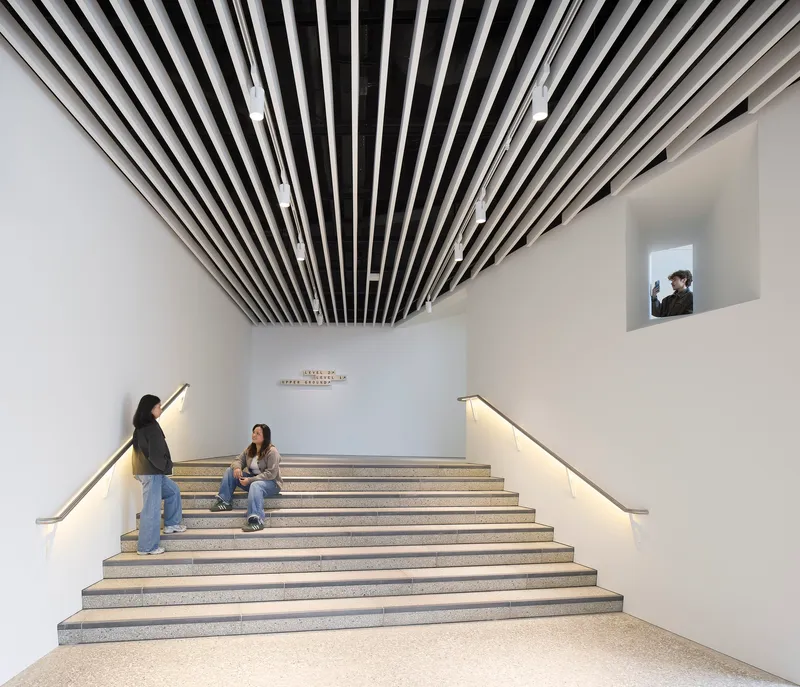 Wide staircase with illuminated handrails beneath radiating slatted ceiling baffles and visitors seated on the steps