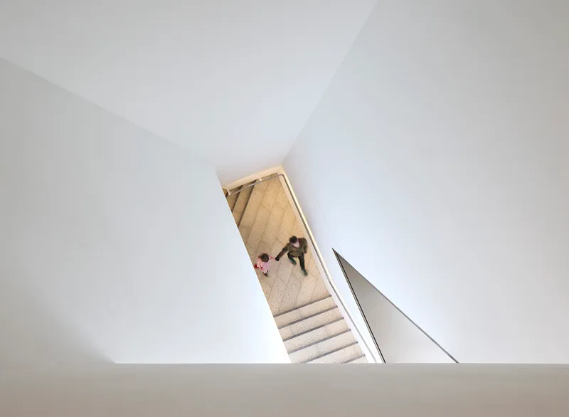 Overhead view of the white stairwell with figures descending the stone treads between angled walls