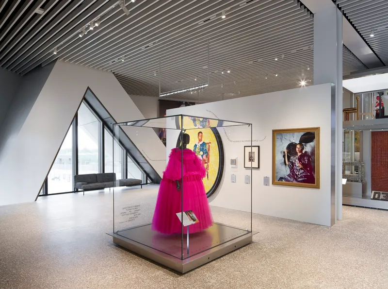 Gallery interior with slatted ceiling and magenta garment in glass vitrine beside angled glazing