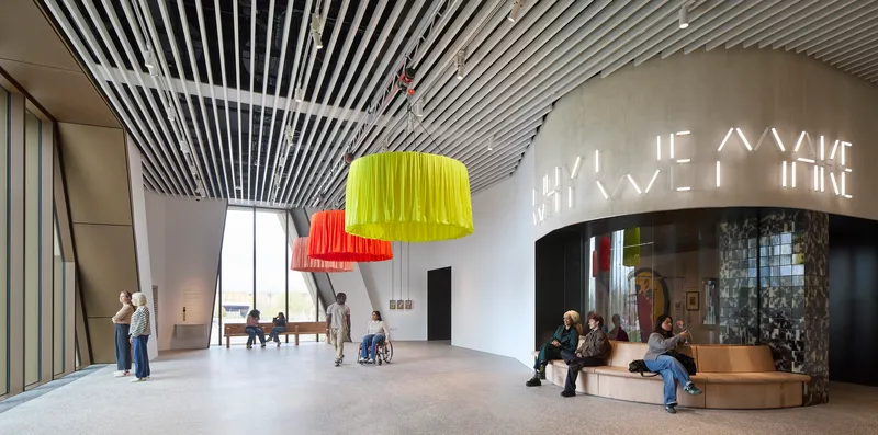 Double-height foyer with yellow and orange pendant lights and visitors beneath curved slatted ceiling