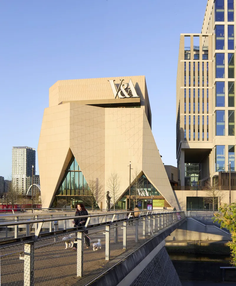 Pedestrian bridge crossing the canal with view toward the faceted facade and adjacent glass tower in afternoon light