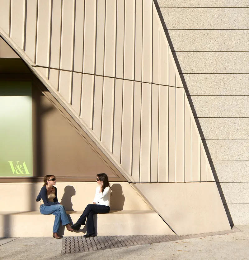 Exterior corner with vertical fluted panels and textured stone where two visitors sit in midday sun