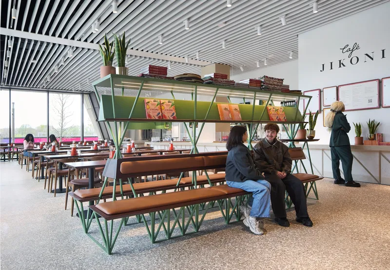 Cafe seating with green metal bench and shelving unit beneath a slatted ceiling with visitors nearby