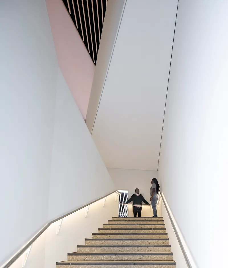 View up the angular stairwell with pink-tinted walls and two figures ascending toward daylight