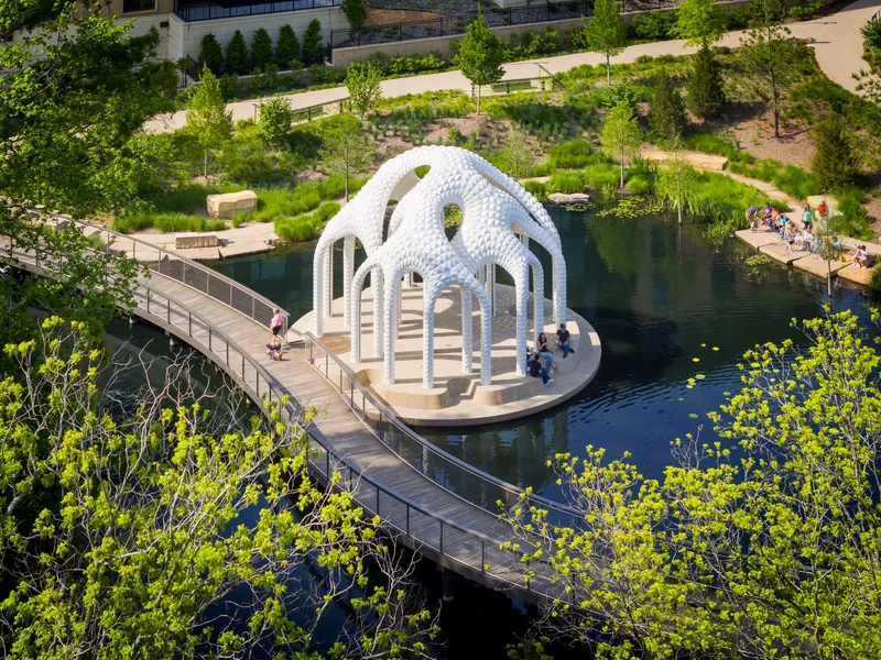 Aerial view of white pavilion with arched columns on circular platform in pond accessed by timber boardwalk