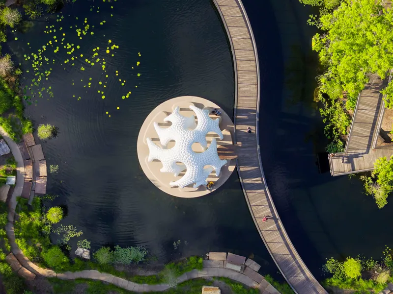 Overhead view of circular pavilion floating in dark pond with lily pads and curving timber walkway