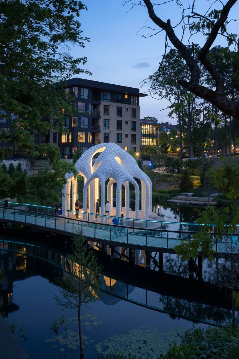 Illuminated pavilion reflected in still pond water at dusk with residential buildings beyond